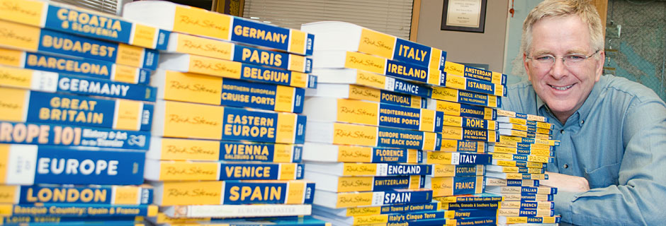 Rick Steves posing next to multiple stacks of guidebooks